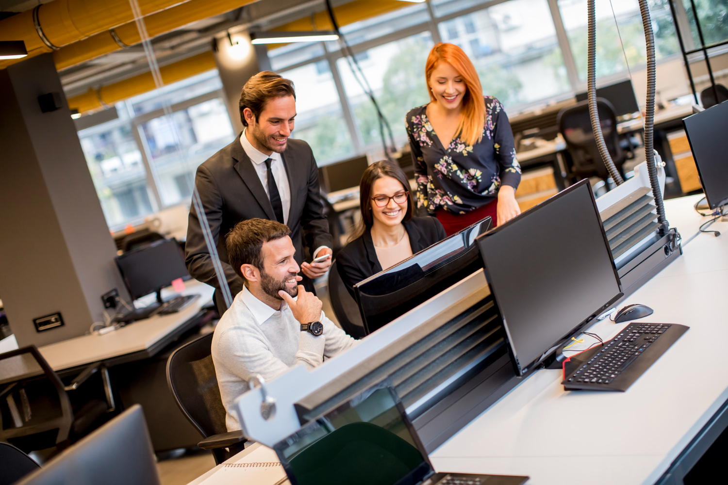 Four people – two men and two women; two are sitting, two are standing, all looking at a computer monitor