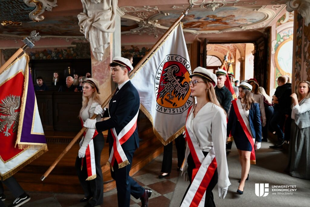 Troje studentów wnosi sztandar Uniwersytetu Ekonomicznego we Wrocławiu do zabytkowej sali.